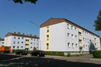 Wohnhäuser mit drei Etagen, Satteldach, Fassade hellgrau und gelb akzentuiert. Grünflächen zwischen Häusern, vorne Straße.