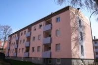 Häuser mit Flachdach, 3 Etagen. Fassade altrosa, Balkonbrüstungen farbich abgesetzt. Davor Grünfläche.