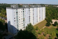 Sanierter Wohnblock, Flachdach, 9 Etagen. Fassade in hellen Blautönen. Loggienbrüstungen mittelblau. Davor Grünfläche mit Bäumen. 