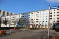 Sanierter Wohnblock, Flachdach, 6 Etagen. Fassade hellblau, Brüstungen Loggien gelb. Vorne gepflasterter Platz mit Brunnen und Bäume. Straße.