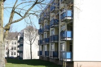 Zwei Wohngebäude, 4 Etagen, blaue Balkone, helle Fassade. Vorne Baum im Winter.