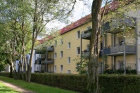 Gelbes Mehrfamilienhaus mit Balkonen, drei Etagen. Vorne sind Hecken und Bäume.