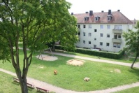 Sandkasten, Schaukel und Wipppferd auf einer Wiese von einem Balkon aus gesehen.
