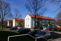 Sanierte Mehrfamilienhäuser mit angestellten Balkonen, 3 Etagen, Satteldach. Vorne Treppe, Grünflächen mit Hecken, Parkplätze.