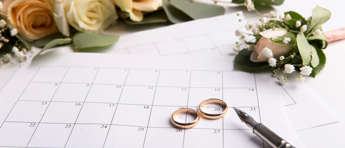 Zwei Eheringe liegen auf einem Kalender, daneben ein Brautstrauß und ein Füllfederhalter Zwei Eheringe liegen auf einem Kalender, daneben ein Brautstrauß und ein Füllfederhalter