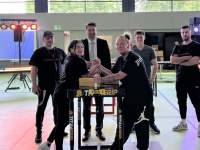 Zwei Personen stehen an einem Tisch und stützen ihre Ellebogen auf. Dahinter befinden sich weiter Personen. 