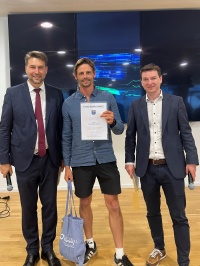 Links  steht ein Mann im Anzug mit Mikrofon, in der Mitte ein Mann in Sportkleidung mit Urkunde und Tüte, rechts ein Mann mit einem Umschlag und Mikrofon