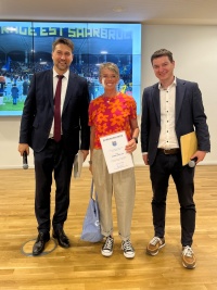 Links  steht ein großer Mann im Anzug mit MIkrofon, in der Mitte eine Frau mit Urkunde und Tüte, rechts ein Mann mit einem Umschlag und Mikrofon