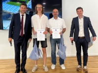 Links  steht ein Mann im Anzug mit Mikrofon, in der Mitte  zwei Männer mit Urkunden und Tüte, rechts ein Mann mit einem Mikrofon