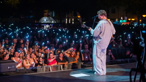 Altstadtfest 2025: Kamrad Ein Sänger steht auf einer Bühne im Scheinwerferlicht. Er hält eine Gitarre, trägt lockere Kleidung und eine Brille. Vor ihm ist ein Mikrofon auf einem Ständer. I