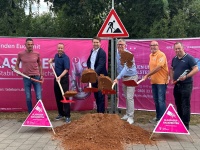 Sechs Männer vor einem magentafarbigen Werbebanner mit Spaten in der Hand