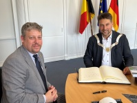 Zwei Männer sitzen an einem Tisch vor einem großen Buch. Im Hintergrund sind die belgische, die europäische und die deutsche Fahne zu sehen.