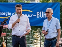 Oberbürgermeister Uwe Conradt und eine weitere Person sprechen auf einer Bühne am Wasser mit Mikrophon.