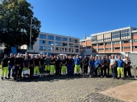 Eine Gruppe von Menschen in Warnkleidung steht auf Pflastersteinen vor zwei Gebäuden