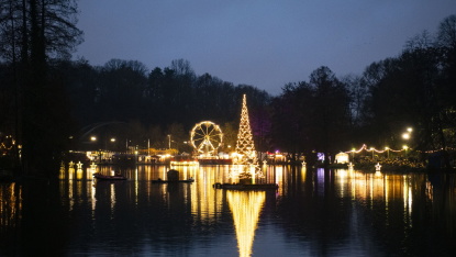 Blick über den See auf den schwimmenden Weihnachtsbaum, Marktlichter und das Riesenrad bei einbrechender Dunkelheit.