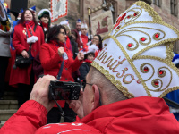 Ein Mann in roter Jacke fotografiert die Szene, während im Vordergrund eine große verzierte Narrenkappe zu sehen ist.