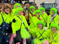 Mehrere Personen in neon-grünen Perücken und leuchtenden Kostümen stehen zusammen vor einem Umzugswagen und lächeln in die Kamera.