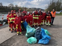 Gruppe von Personen in roten Einsatzkleidungen stehen am Ufer eines Flusses neben gesammelten Müllsäcken